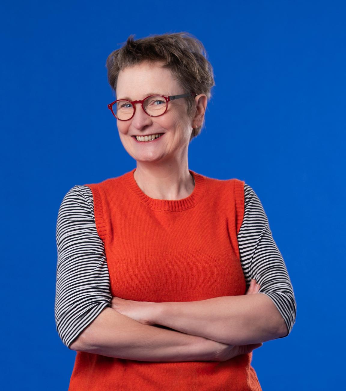 A smiling woman with short dark hair wears red-framed glasses, a grey top and red vest and is pictured against a bright blue background