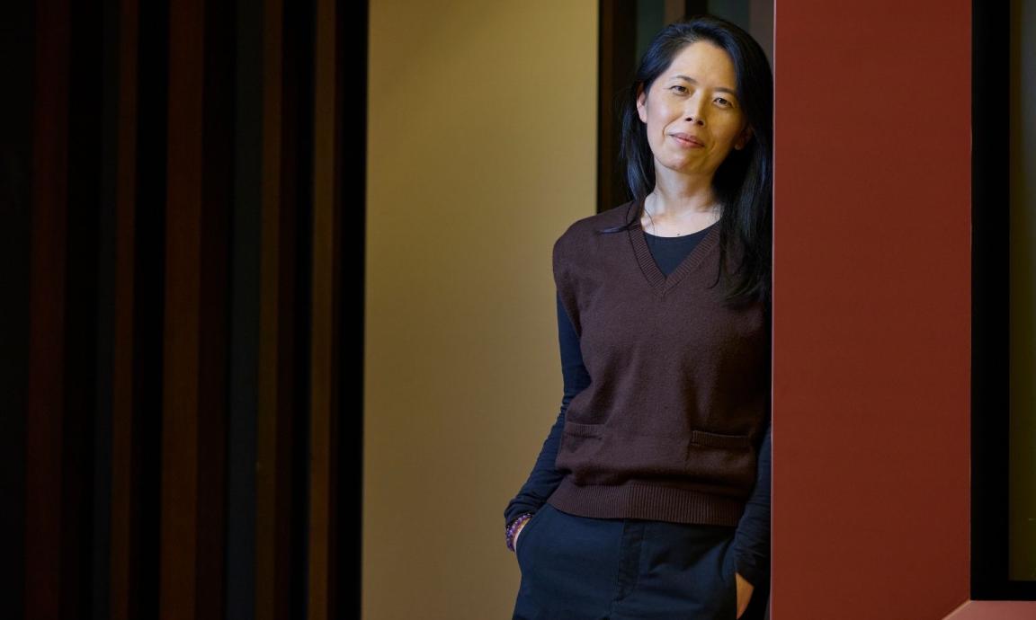 A woman with long dark hair stands leaning against a wall, smiling gently at the camera. She wears a brown V-neck sweater over a black shirt and dark pants, with her hands in her pockets.