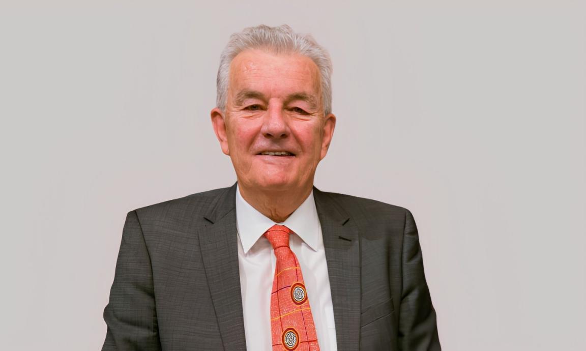 A smiling man with grey hair wearing a grey suit, white shirt and a red patterned tie with circular emblems. He is photographed against a plain light gray background.