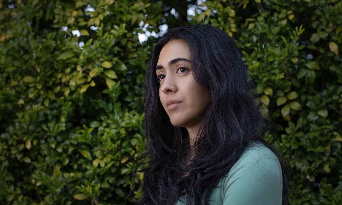 A woman with long dark hair wearing a green shirt is pictured in front of green shrubbery