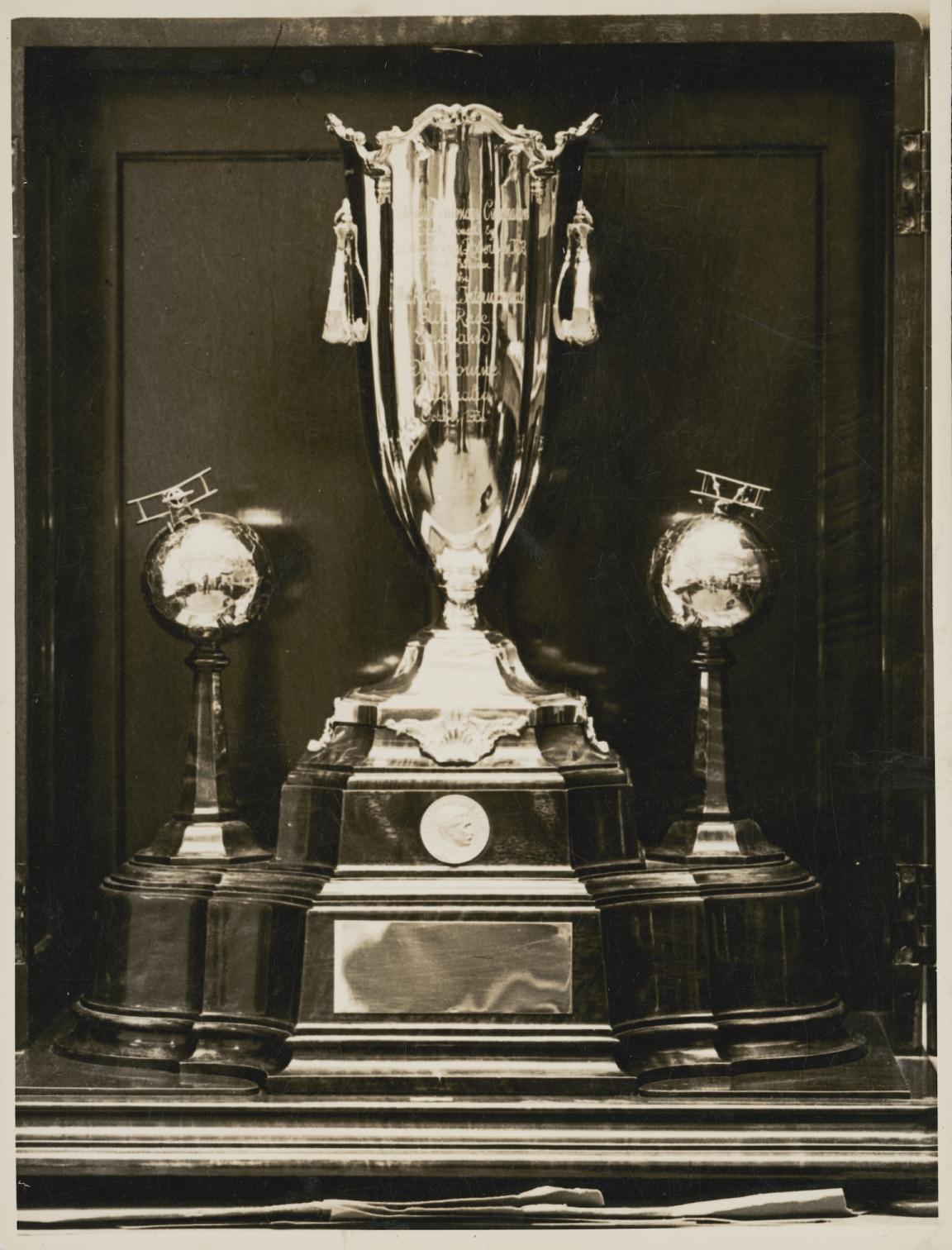 Black and white photo of a large trophy with a wide base from which extends a large cup and two small globes with planes on them