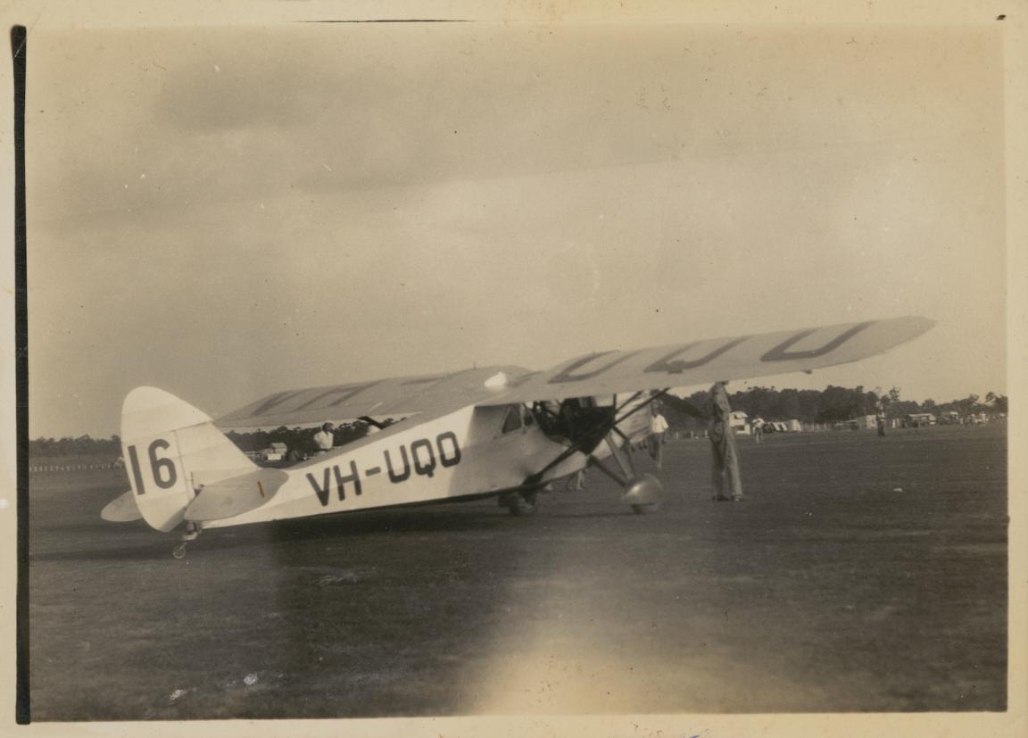 Sepia-toned photo of a smal plane parked in a field, with the pilot standing in front of it