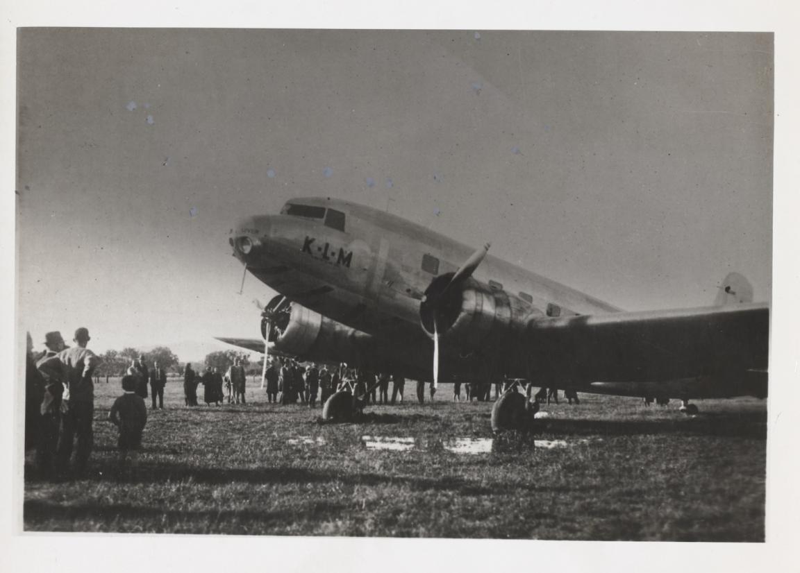 Black and white photo of a plane bogged in a racecourse, with serveral people standing around it