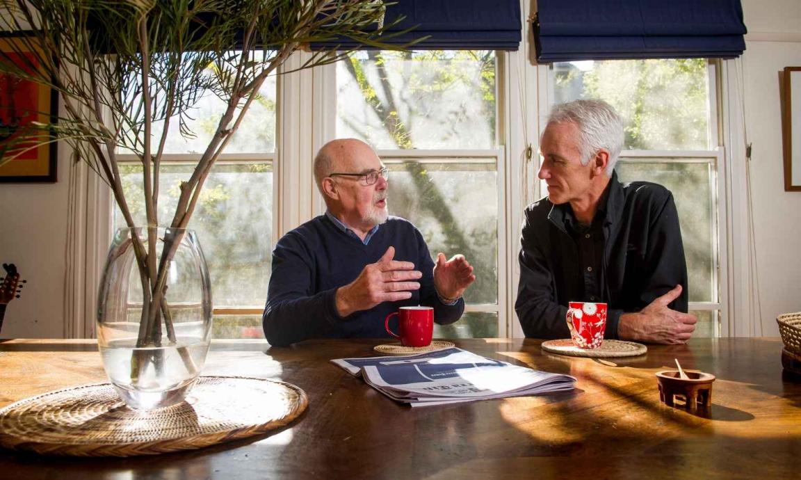 Two men engaged in conversation at a wooden table near a window. The man on the left, wearing glasses and a dark sweater, is gesturing as he speaks. The man on the right, with gray hair and a dark shirt, listens attentively. Both have mugs in front of them, and a vase with branches sits on the table. Natural light streams in from the window behind them.