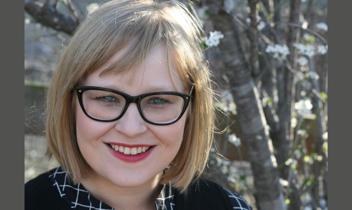 A smiling woman with short blonde hair and black-rimmed glasses, wearing a black and white checkered top. She has a nose ring and is outdoors with a blurred background of flowering trees.
