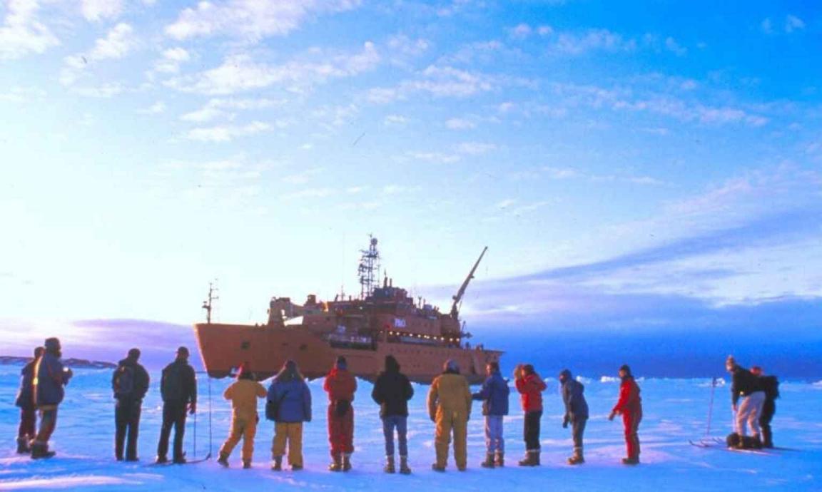A group of people in colorful winter gear stand on a vast expanse of snow and ice, looking towards a large orange icebreaker ship in the distance