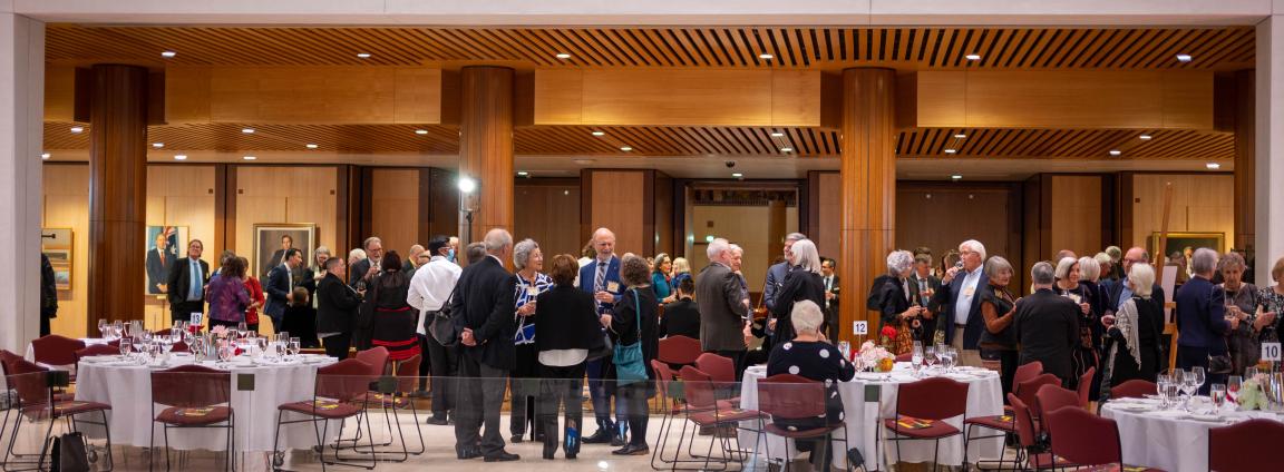 A group of adults in formal attire mingle and converse in a spacious, wood-paneled event hall with round tables set for dining.