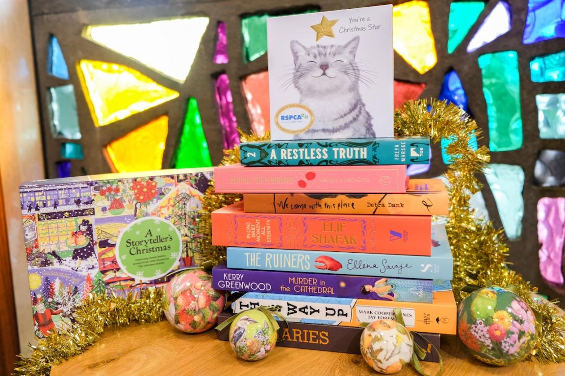 A stack of books sitting on a table in front of a colourful window and surrounded by Christmas decorations.