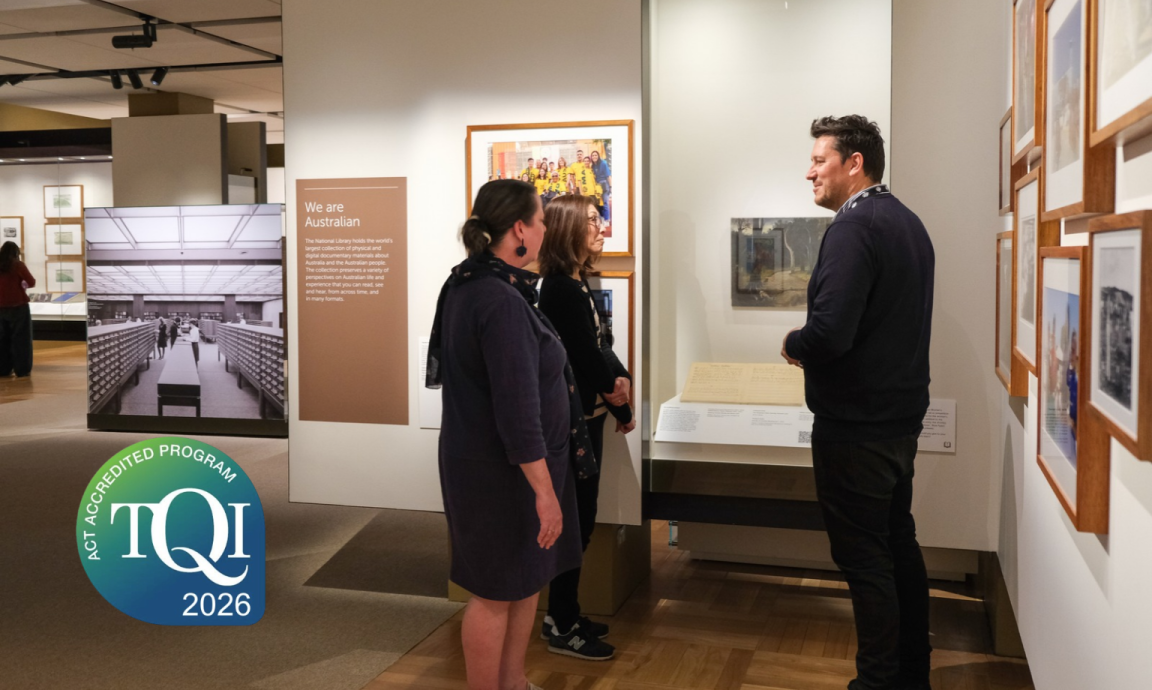 Three people are viewing exhibits in a museum gallery. A man stands facing two women, appearing to explain something about displayed artwork and documents. A large informational panel titled 'We are Australian' is visible, along with framed pictures on the walls and a display case. A 'TQI ACT Accredited Program 2026' logo is in the lower-left corner.