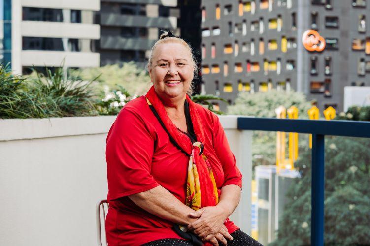 A smiling woman with her hair tied up sits on a chair on a balcony, wearing a red top and a red and yellow scarf.