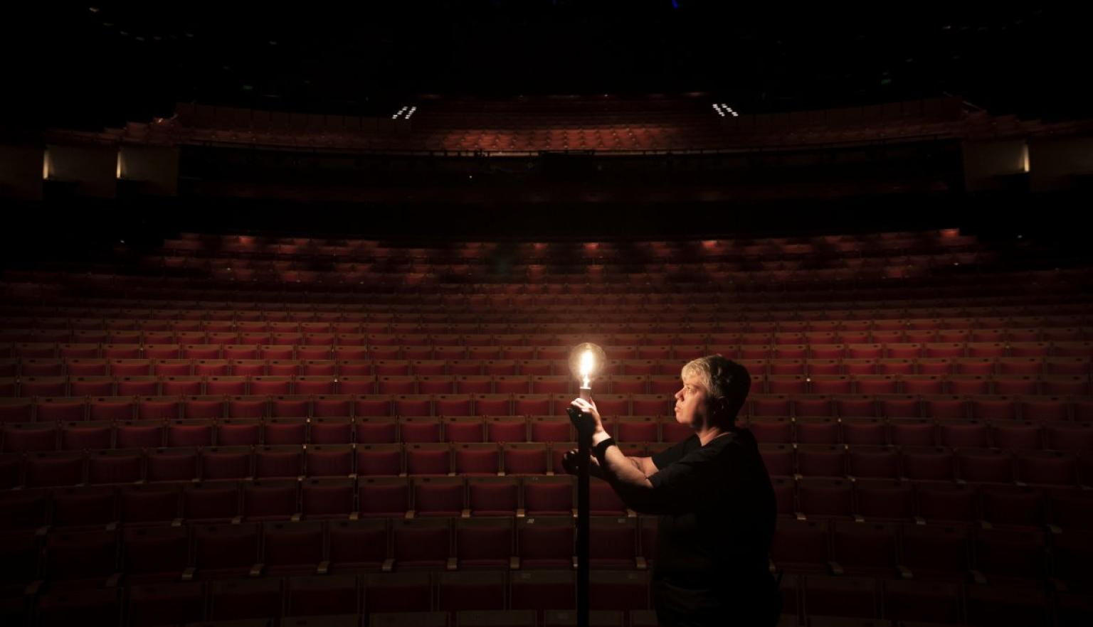 A person stands in an empty darkened theatre, illuminated by a single light bulb.
