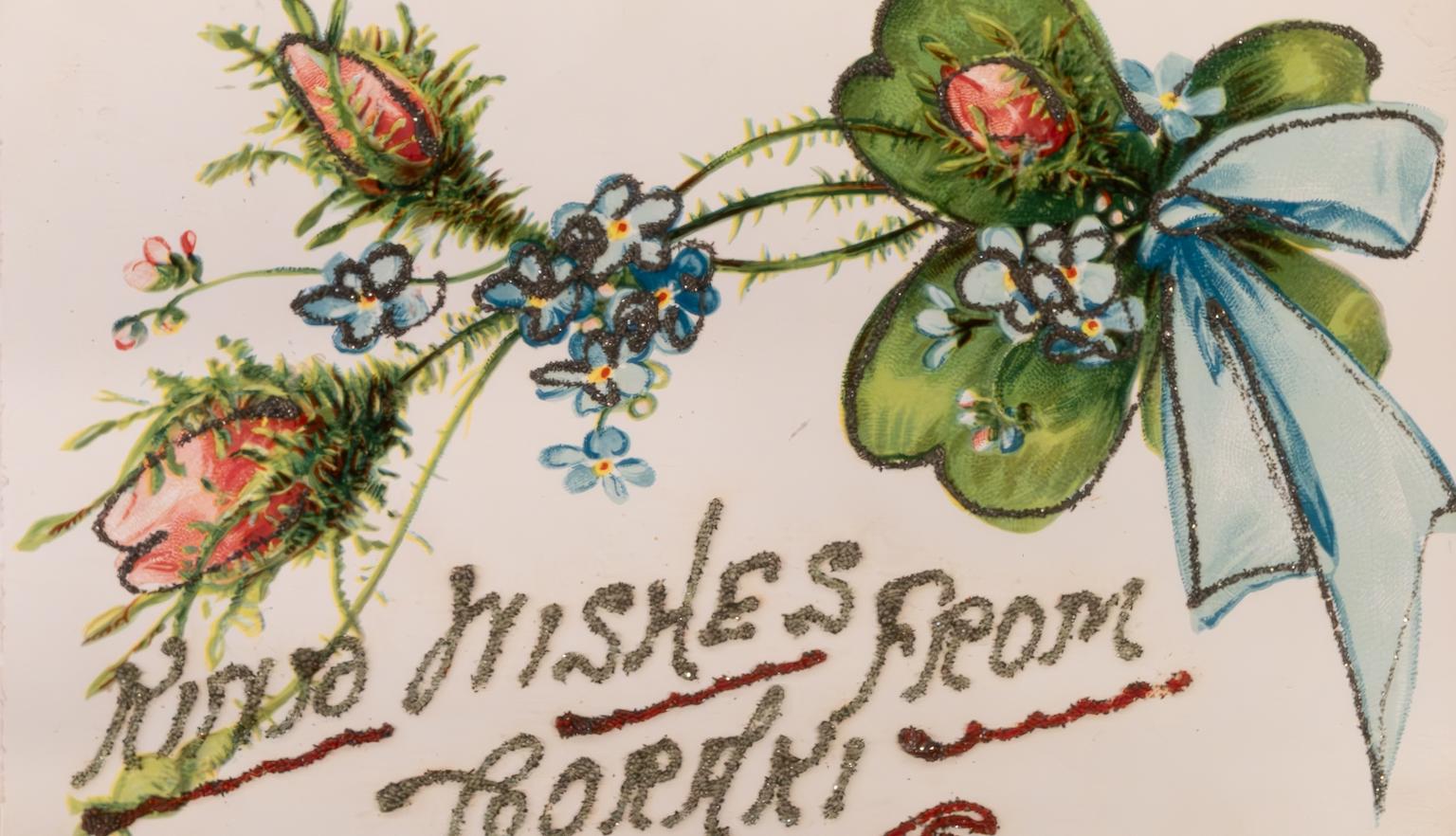Front of an old postcard featuring a design with glitter of flowers and a ribbon with glitter text reading 'Xmas wishes from Coraki'.