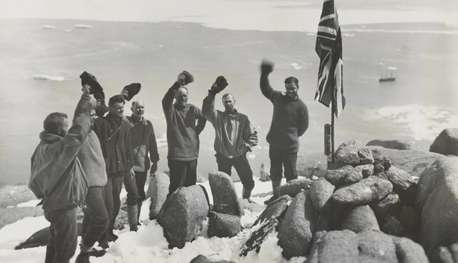 Group of explorers standing on the top of a snowing cliff overlooking a harbour, waving near a Union Jack flag