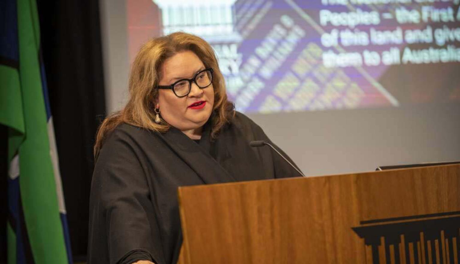 A person with long brown hair and glasses stands behind a wooden lectern, speaking into a microphone. The background features a screen displaying partially visible text and graphics. To the left, green and blue from the flag of the Torres Strait Islands is visible.