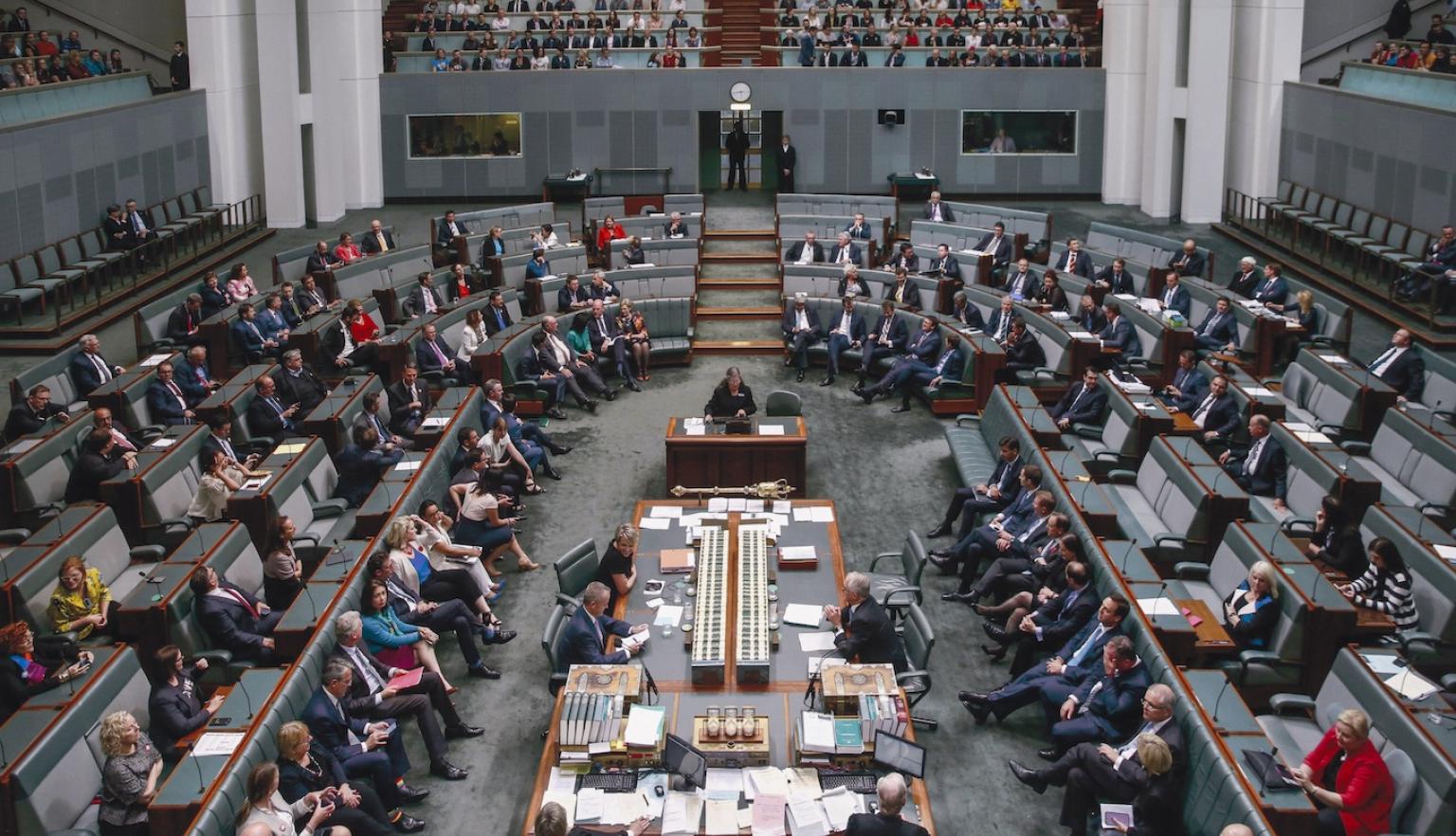 View of the Australian House of Representatives in Parliament House from slightly above. Members of the House of Representatives sit throughout the room, and members of the public sit in allocated areas