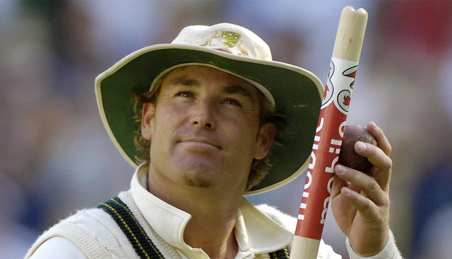 A man in cricket whites and a hat holding a cricket stump and looking upwards.
