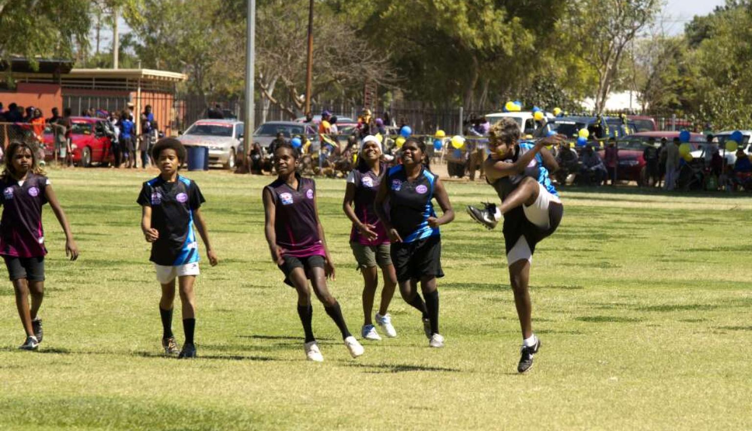 Six women smiling as they play football. One has just kicked the ball.