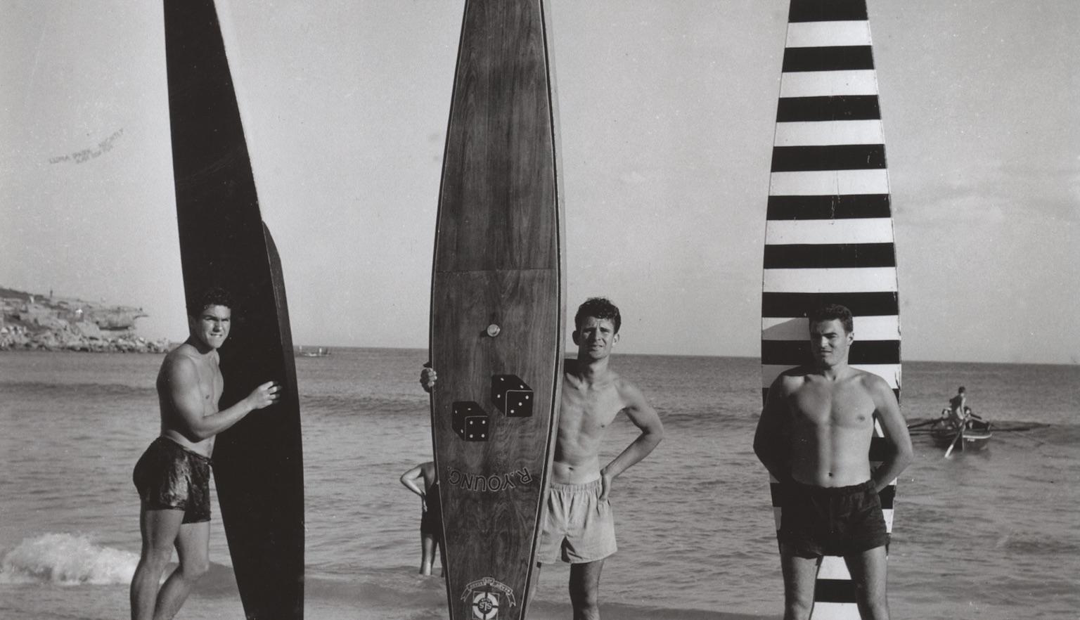Black and white photo showing three men in swimwear holding up long boards on the sand at a beach