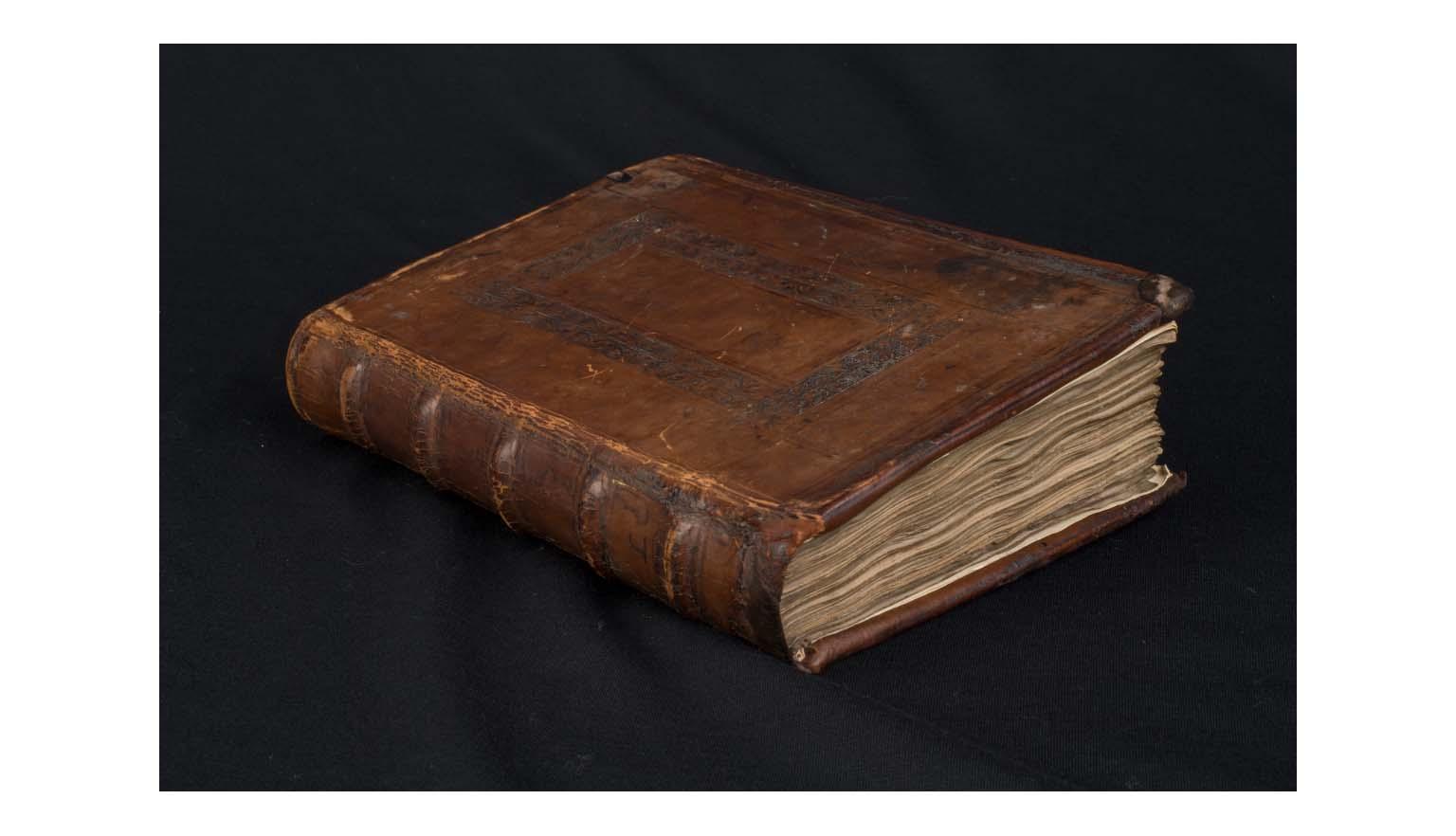 A closed book. It is sitting on a black velvet cloth. The book is bound in brown leather. The front cover and spine are carved and moulded. The front cover is very scratched and worn.