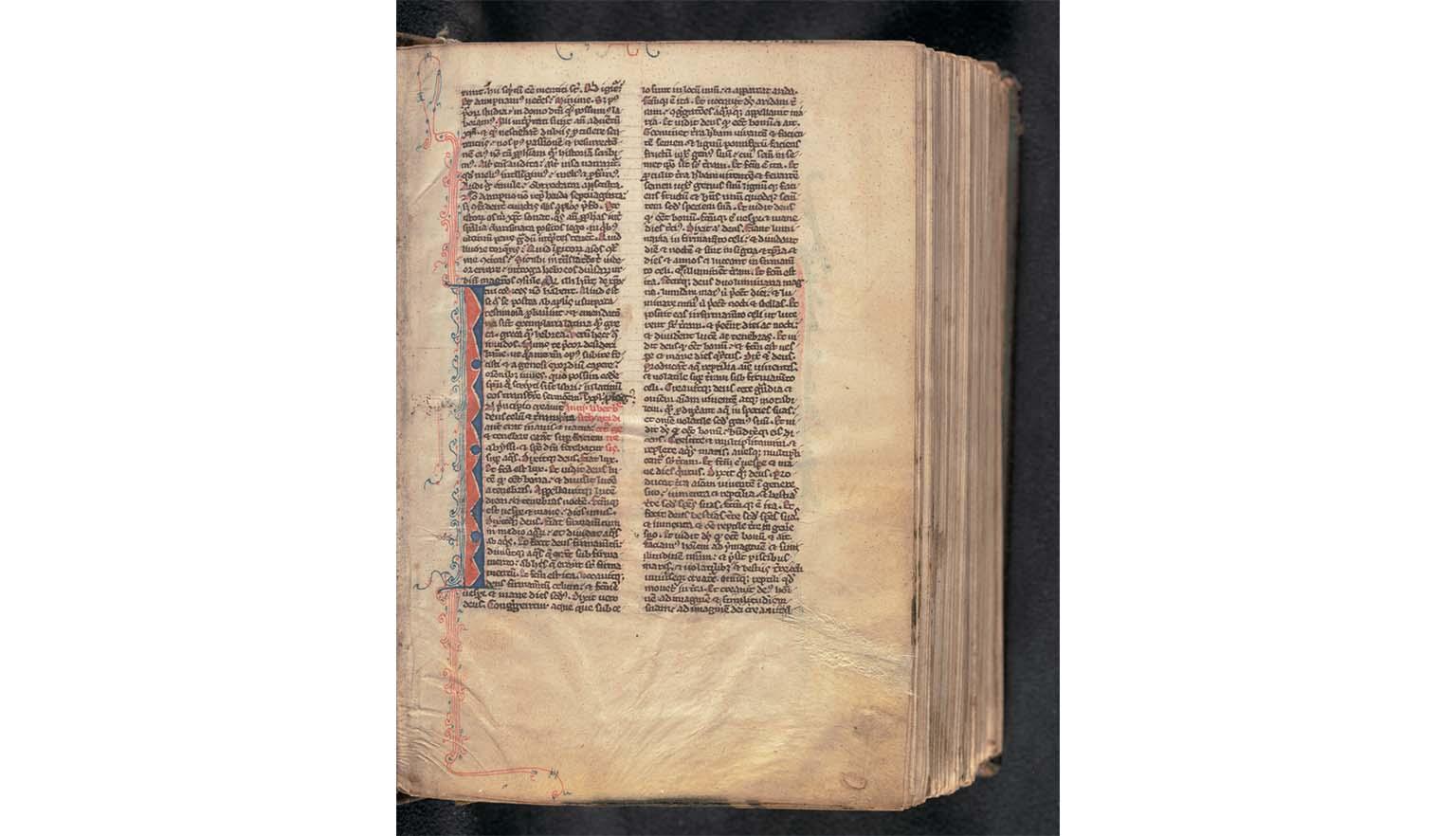 A richly decorated handwritten page from an old bible. It is written in Latin. The text is in black ink. In the margins of the page are blue and red decorations