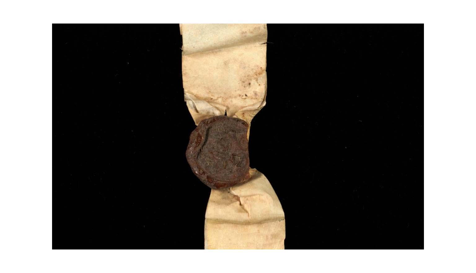 A close up of a red wax seal attached to a strip of paper. The seal pictures a crest but it is very difficult to make out due to age.