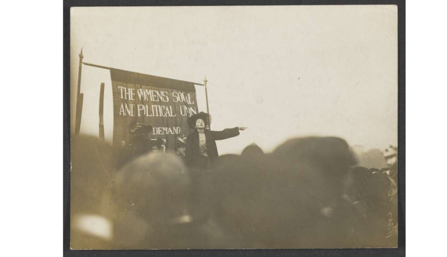 A woman stands on a platform in front of a banner that reads "THE WOMEN'S SOCIAL AND POLITICAL UNION."