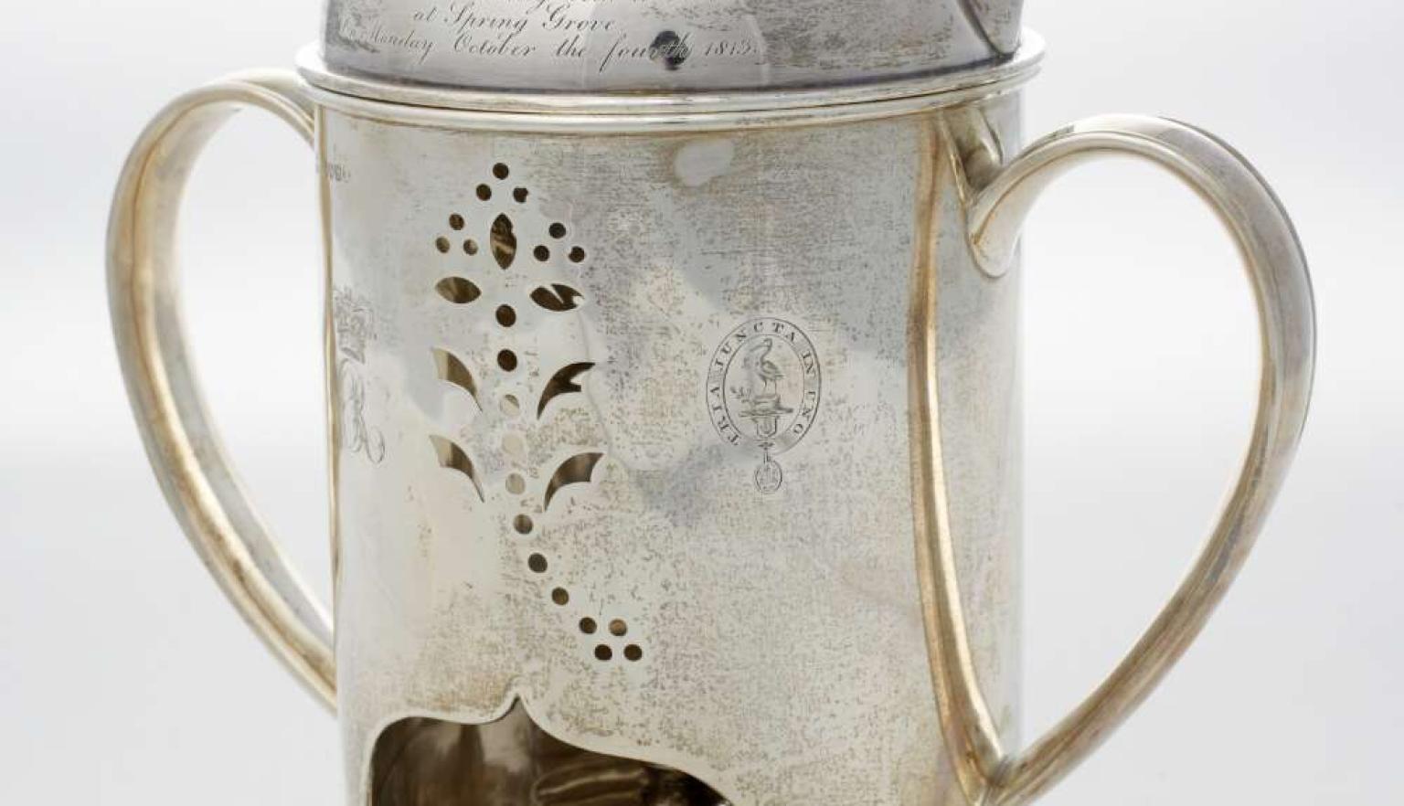 A silver polished kettle with ornate cutouts and inscription and printed crest. It has a tealight underneath.