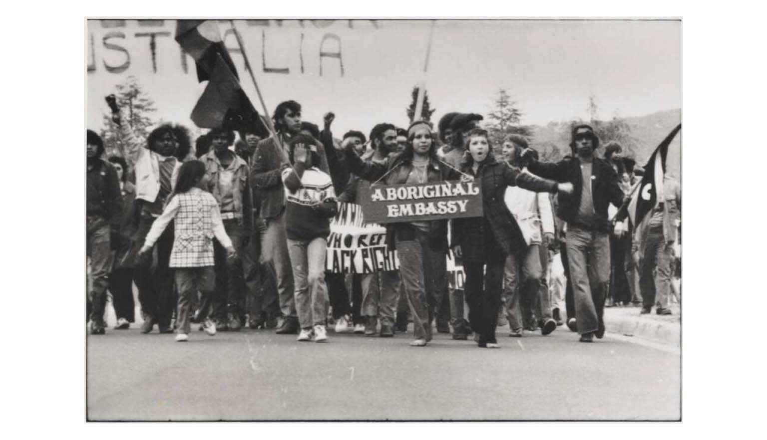 A black and white photo of a large group of people marching along a street, holding large flags and a sign saying 'Aboriginal Embassy'.