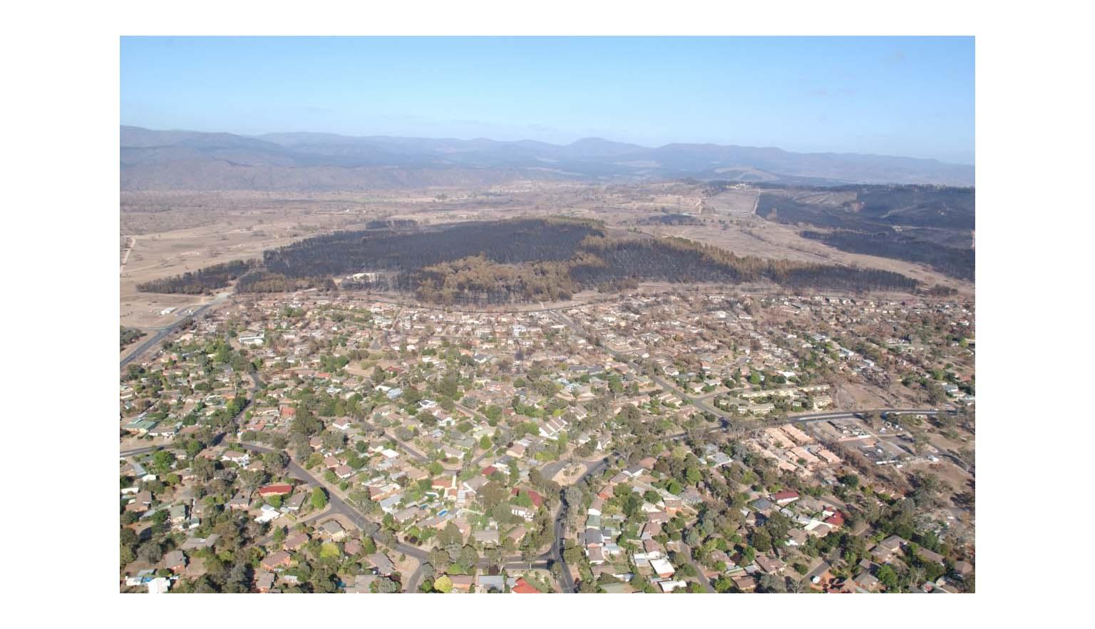 An aerial shot of a city suburb. A hill surrounding the suburb is scarred black from a large fire.