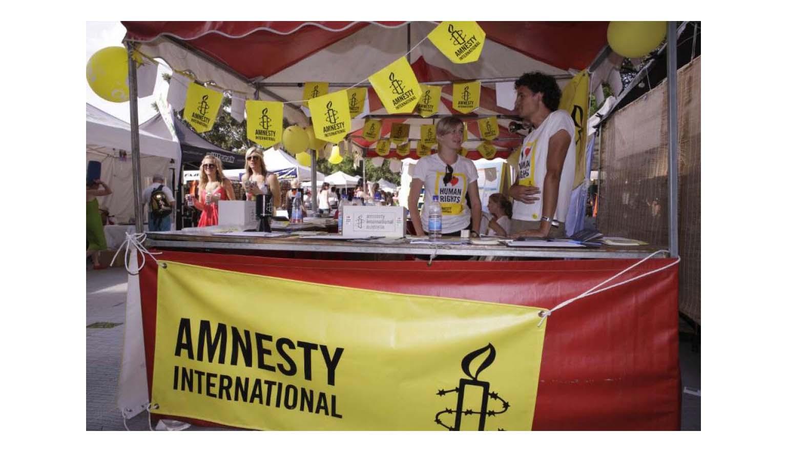 A photo of 2 people talking inside a market stall decorated with red and yellow signage reading 'Amnesty international'.