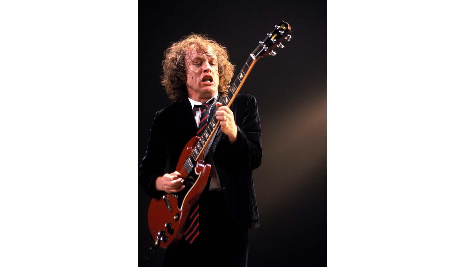A man with long curly hair plays a guitar with great energy. He is very sweaty. The guitar is electric and has a red body. He is wearing a school uniform with a black blazer and a red and black striped tie