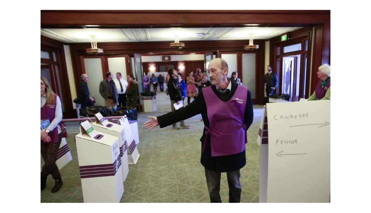 A photo of a man in a purple apron directing people toward ballot boxes.