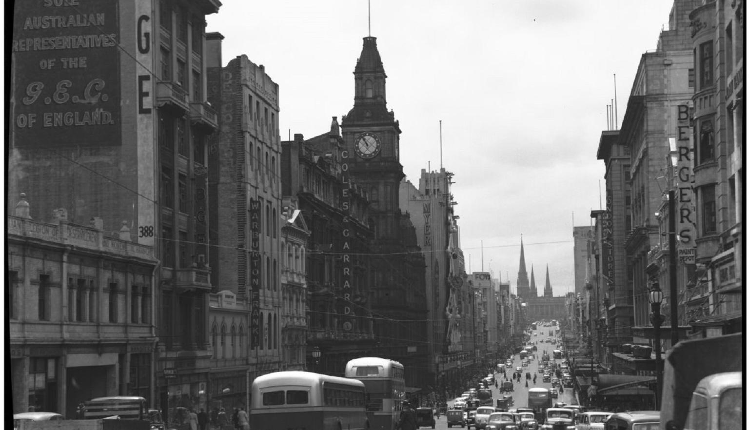 A black-and-white photograph showing Bourke Street, Melbourne, in the mid-20th century, with buses, cars, and pedestrians. The street is flanked by tall buildings with prominent signage, and a clock tower is visible.