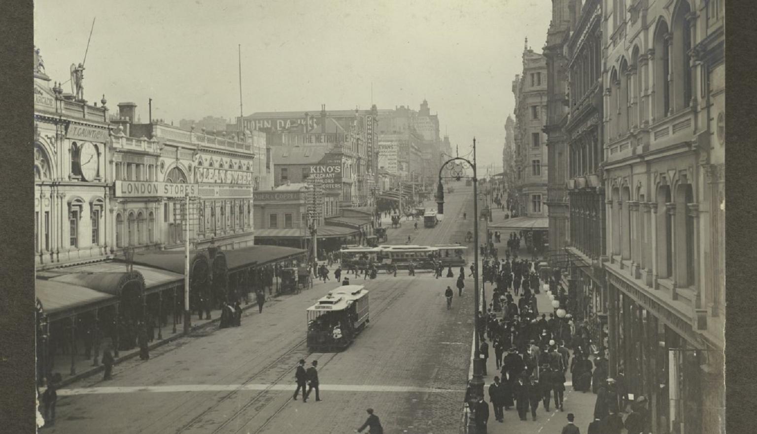 A black-and-white photograph of early 20th-century Bourke Street, Melbourne, with trams, pedestrians, and people on bicycles. The street is lined with ornate building facades, and a crowd walks along the footpath.