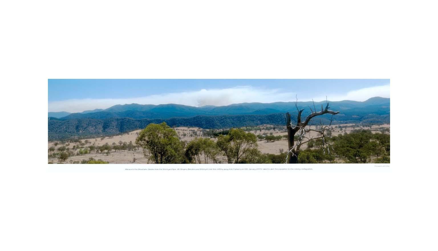 A wide panorama of a mountain range during the day. In the centre of the distant range, a smudge of smoke can be seen rising into the sky