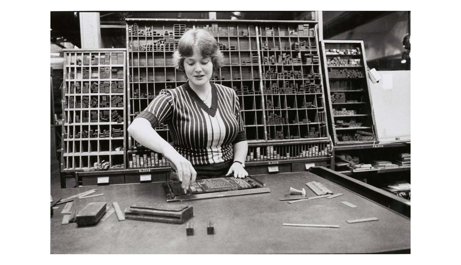 A black and white image of a woman at a desk setting type face into a frame. Behind her is a large rack full of letters