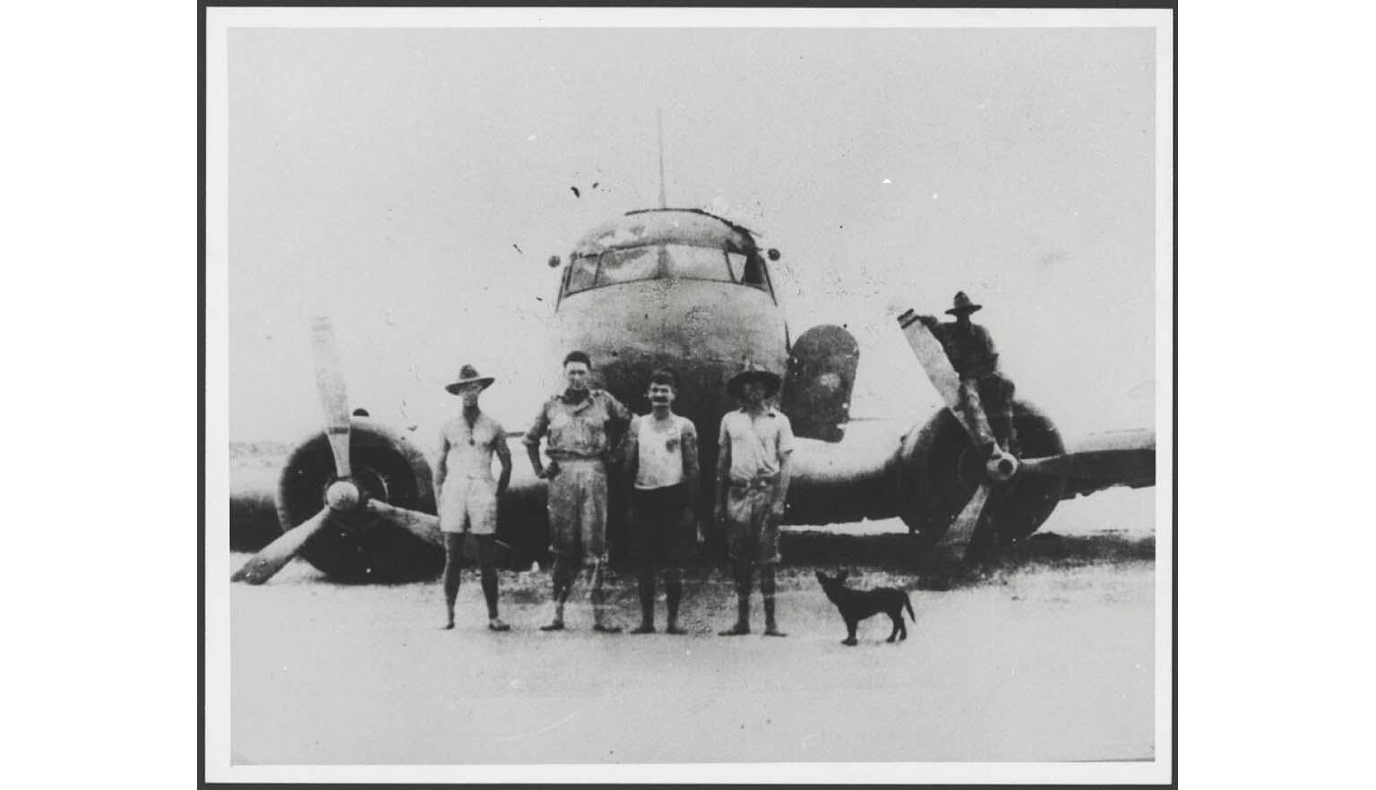black and white photograph of four men and a dog standing in front of a plane
