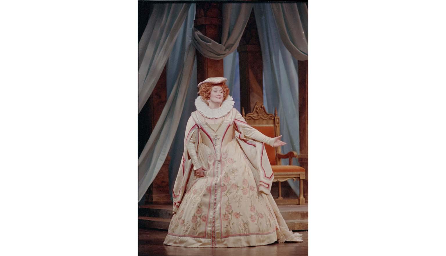 A picture of a woman on stage mid-performance. She is dressed in a large old-fashioned gown with ruffles. Behind her is a throne and heavy curtains