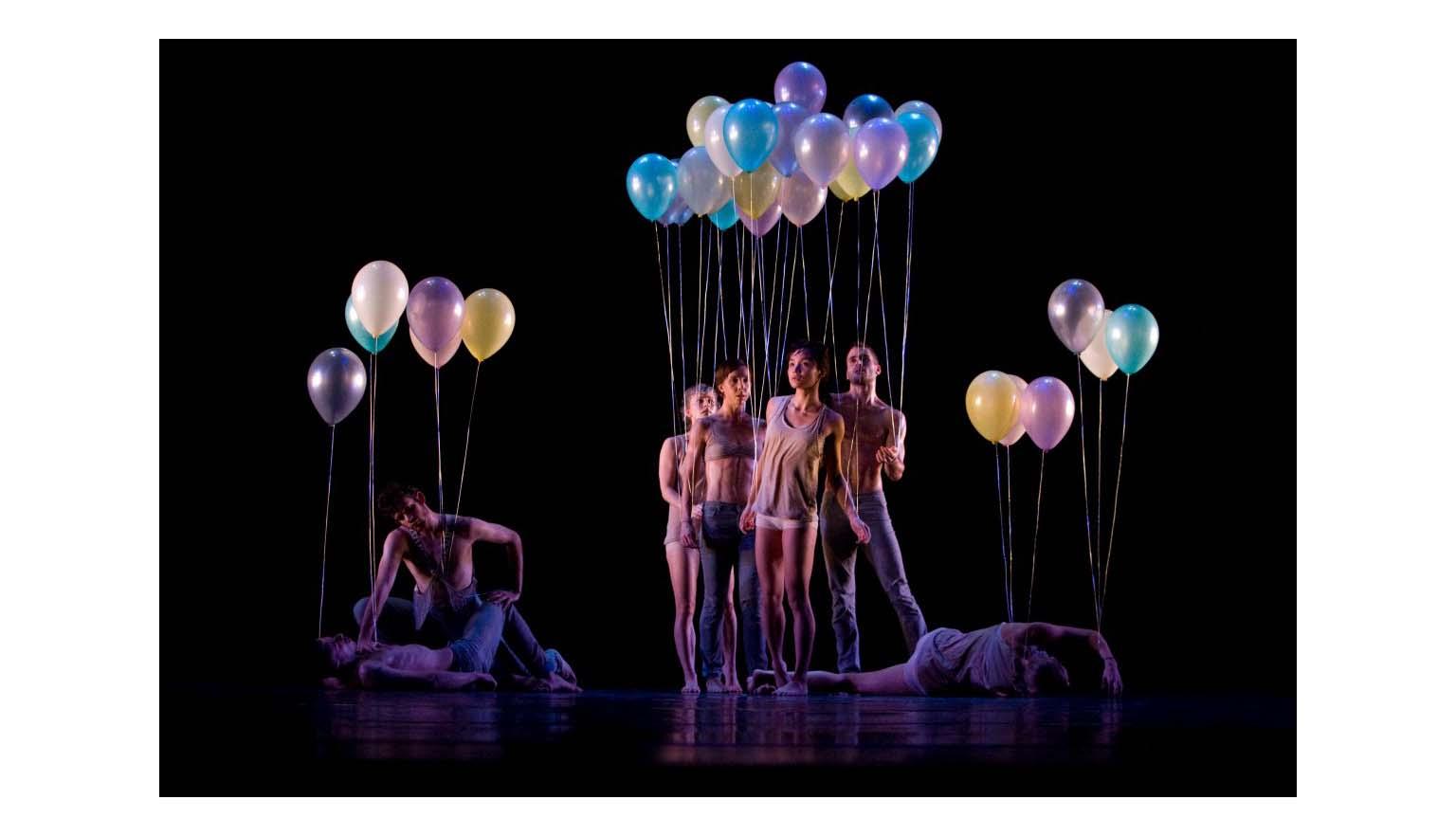 A troupe of dancers illuminated on a blacked out stage. They are holding colourful balloons. Some actors are laying on the floor, most are standing.