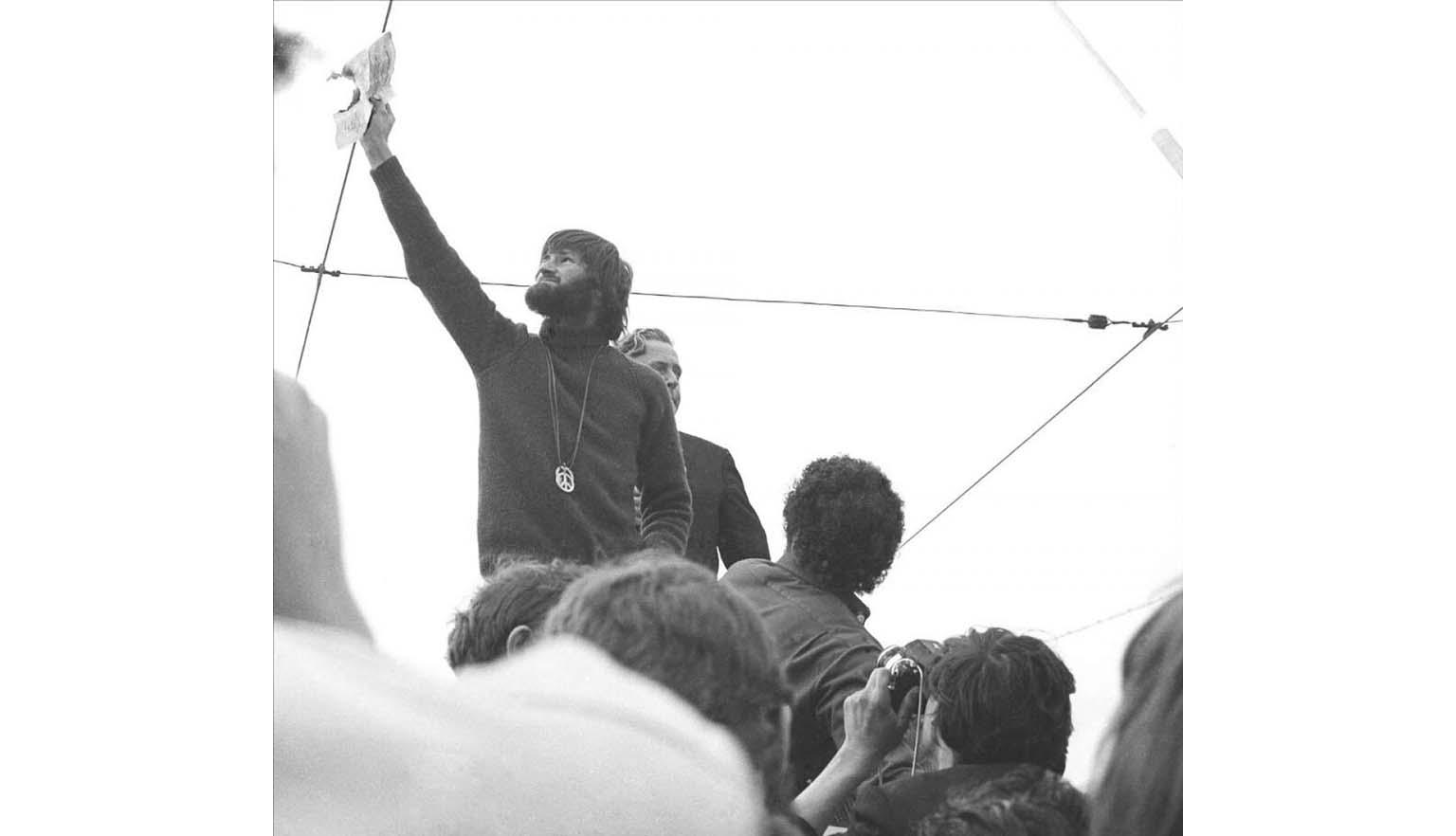A black and white photo of a bearded man with collar length hair and a large metal peace medallion hanging down his front, holds a burning piece of paper up high, while several people watch and take photos.