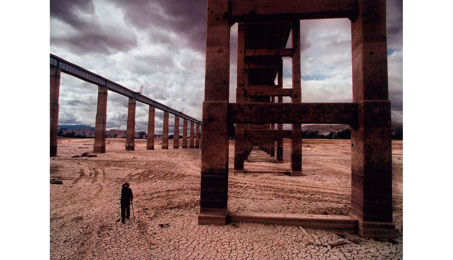 A man with a walking stick walks below the pylons of a huge bridge that have surfaced after a drought. The river bottom is cracked and crazed with the drying of the mud.
