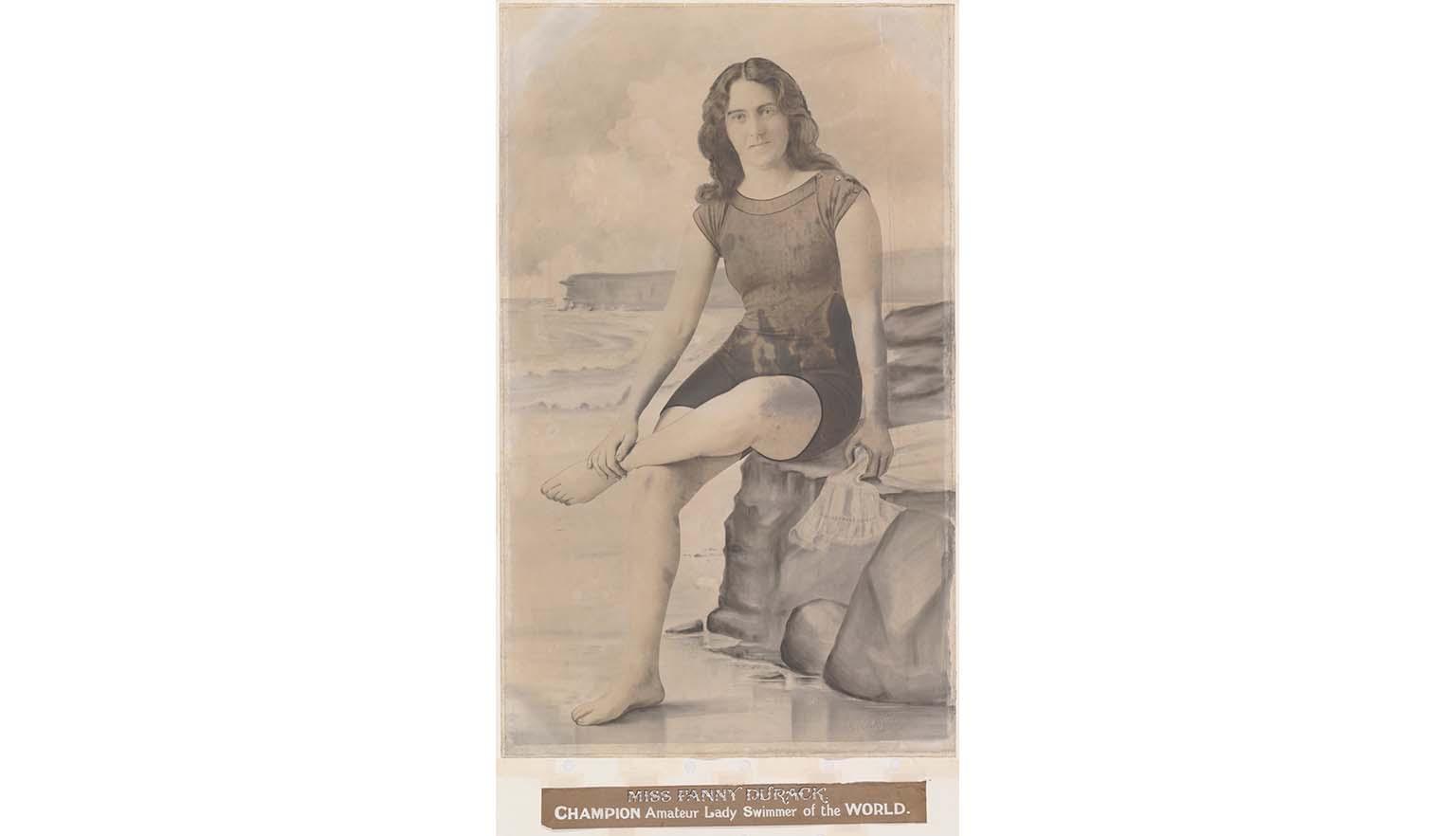 A sepia toned photograph of a woman sitting on a rock. She is wearing an old fashioned swim suit. She is sitting crossed legged. Beneath her is a caption saying 'MISS FANNY DURACK' Champion Amateur Lady Swimmer of the World'