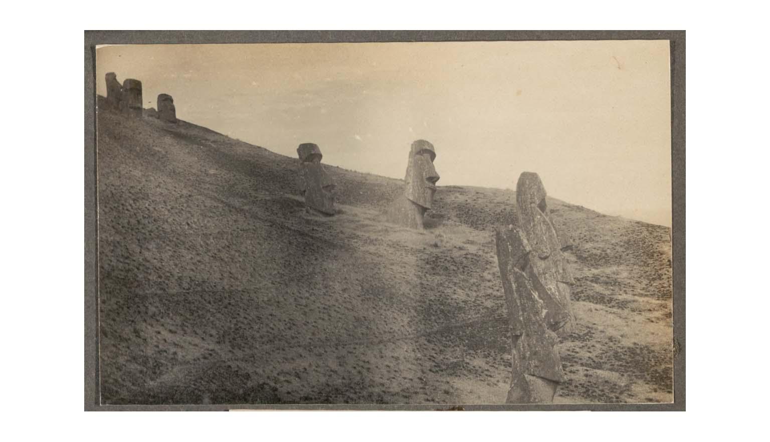 A sepia toned photograph of seven enormous stone carved heads, Moai, dotted along a barren cliff face. The statues are on various angles and have succumb to varying levels of