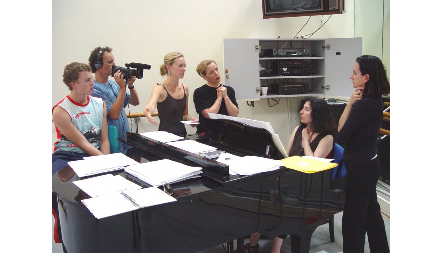 Four women and two men stand around a black baby grand piano in a studio. One man is filming the pianist and is wearing headphones. The group are mid-conversation.