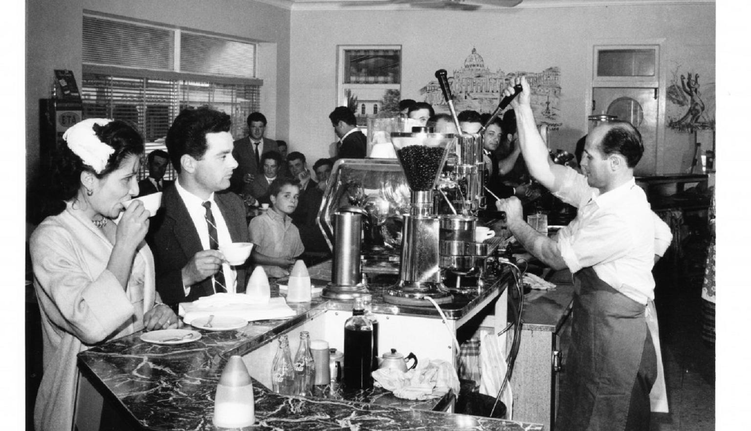 A black-and-white photograph of a busy café scene, with a man operating an espresso machine behind the counter. Customers stand or sit at the counter, drinking coffee.