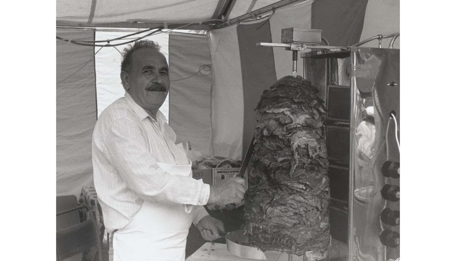 A man with a dark moustache and a white apron is carving meat from a revolving pile to be used for kebabs.