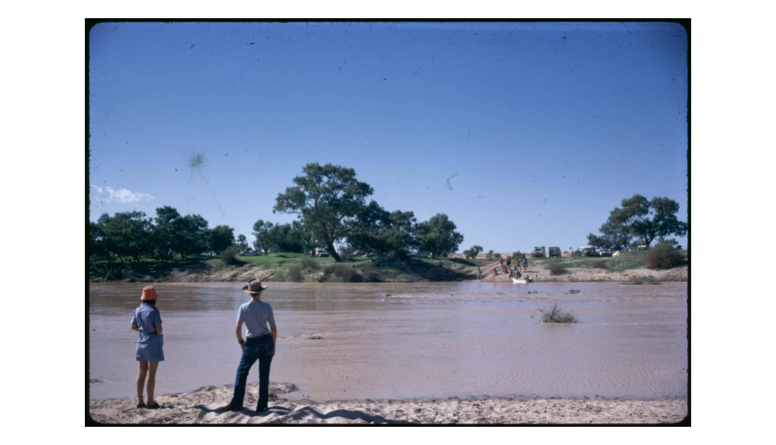 Two people stand on the bank of a swollen river in full flood. The water is turgid and brown. In the distance, a group of people stand on the opposite bank.