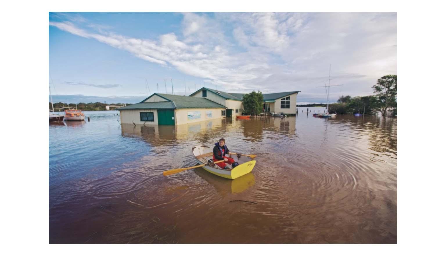 A building is partially submerged by flood water. A man in a dinghy is rowing towards the structure.