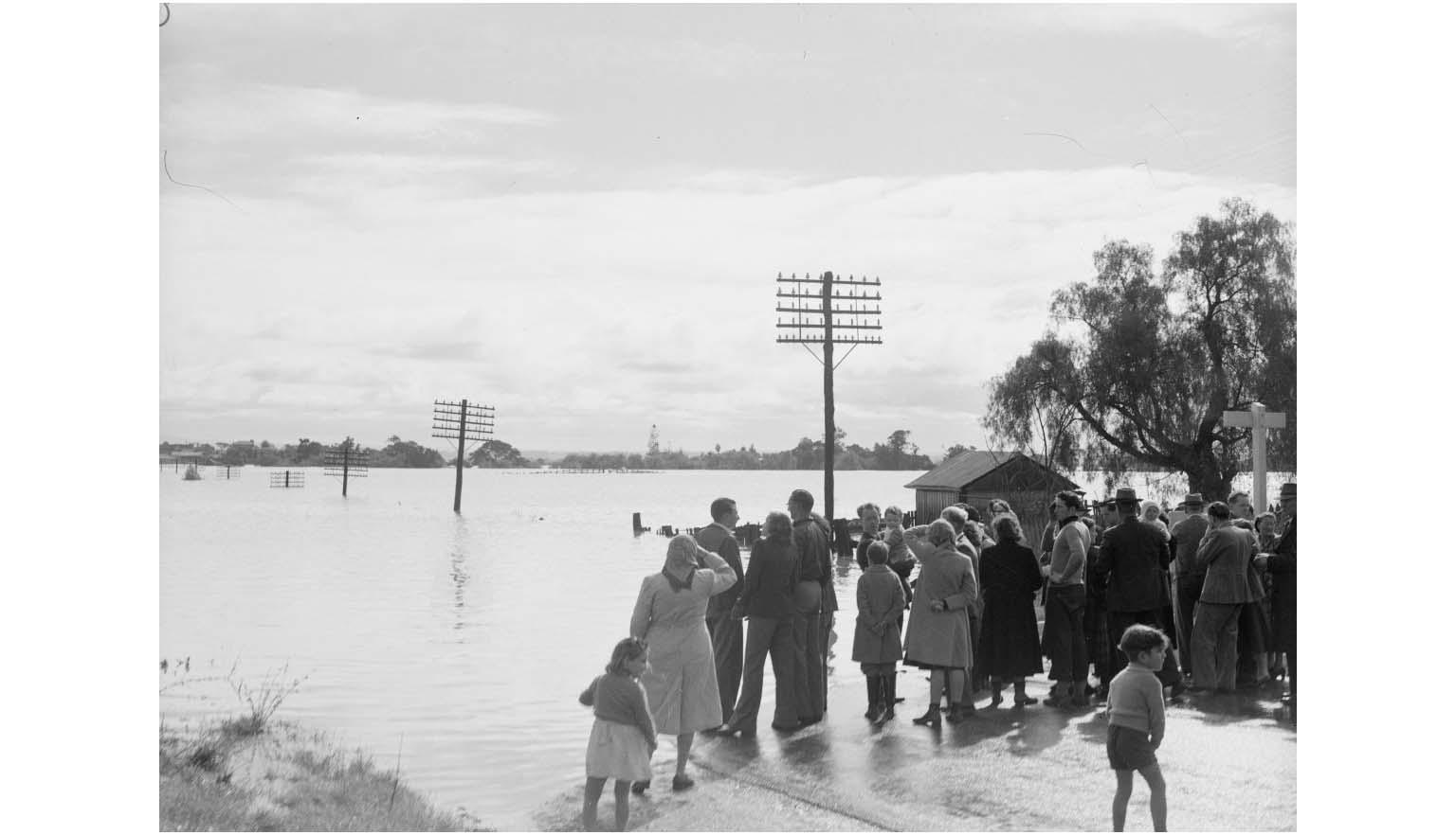 A large group of people stand on a road that is cut off by a swollen flooded river. Telegraph poles are submerged up to the head and trail off into the water.