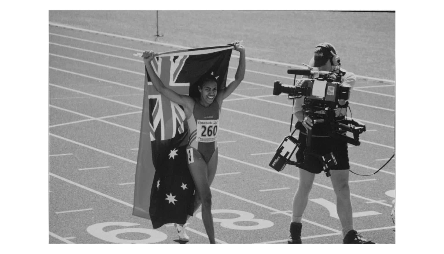 Black and white photo of Cathy Freeman carrying Australian and Indigenous flags with a cameraman filming alongside her